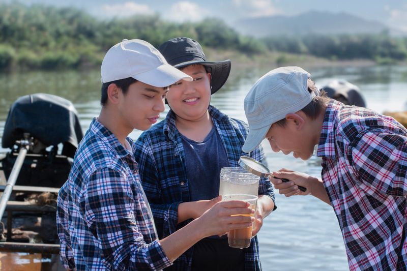Asian boys in plaid shirt hold transparent plastic bottles and  cup which had water from river in front of them inside, they are studying water, environment, river, insects, particles and eco effect.