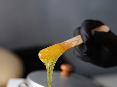 Golden wax dripping from a wooden spatula held by a gloved hand.