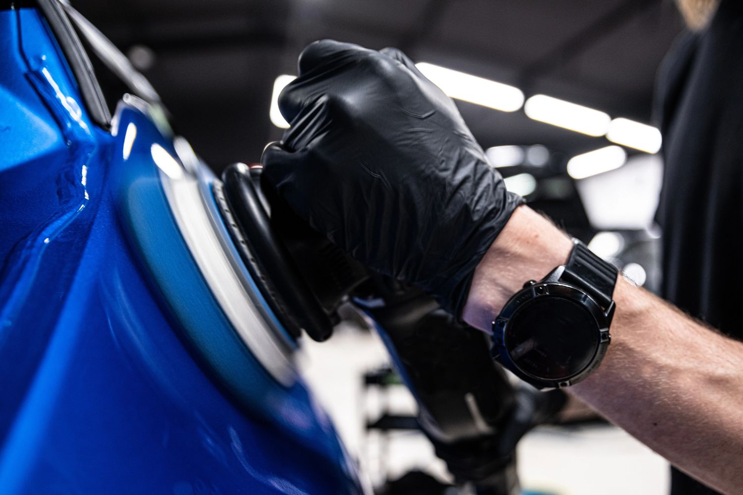 Person wearing black gloves polishing a blue car with a buffer.