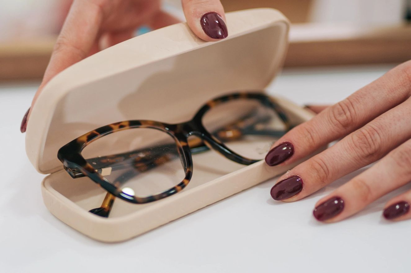 Tortoiseshell glasses in a beige case held by hands with dark nail polish.