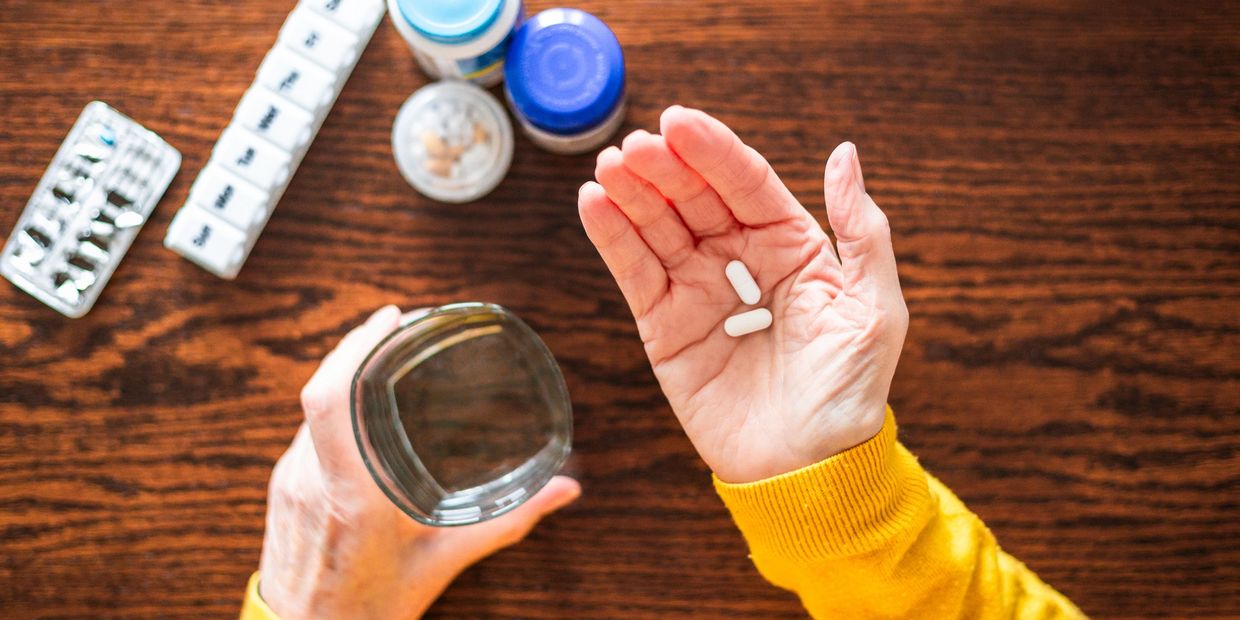 Person holding two white pills and a glass of water over a wooden table with medications.