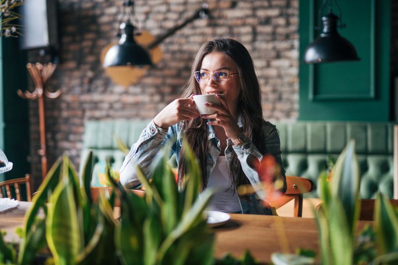 A young beautiful girl is sitting in a coffee shop and enjoying her coffee. In the foreground is a plant.