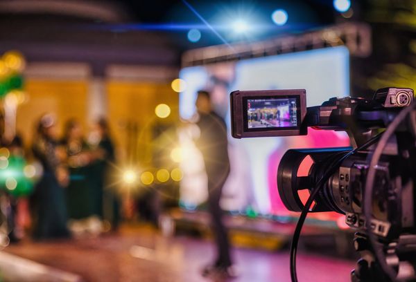 Professional camera recording a blurred event with people and stage lighting.