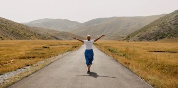 A woman walks barefoot on a long road with arms outstretched in a mountainous landscape.