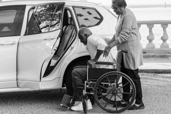 Woman helping man from wheelchair to car.