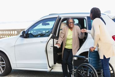 A woman helps an elderly lady out of a car near a wheelchair.