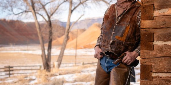 Cowboy standing by a wooden building in a desert landscape.