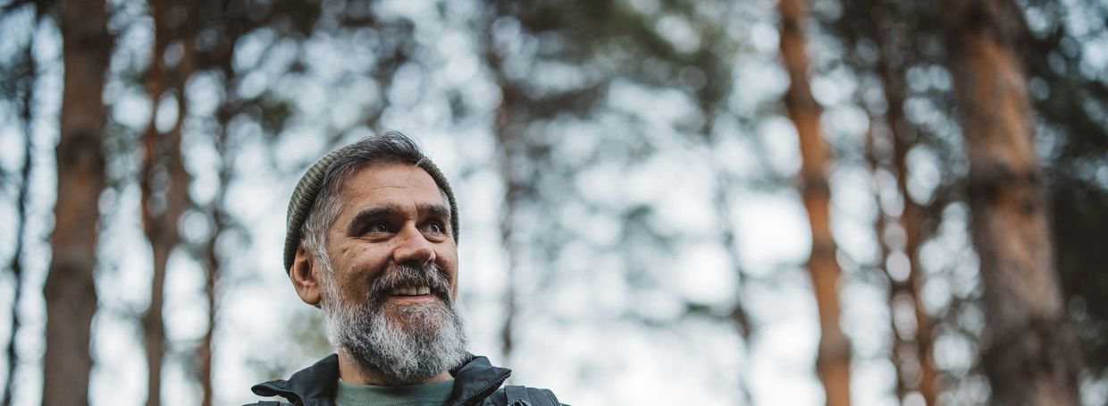 Smiling man with gray beard and beanie outdoors in a forest.