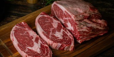 Two raw ribeye steaks and a larger piece of ribeye on a wooden cutting board.