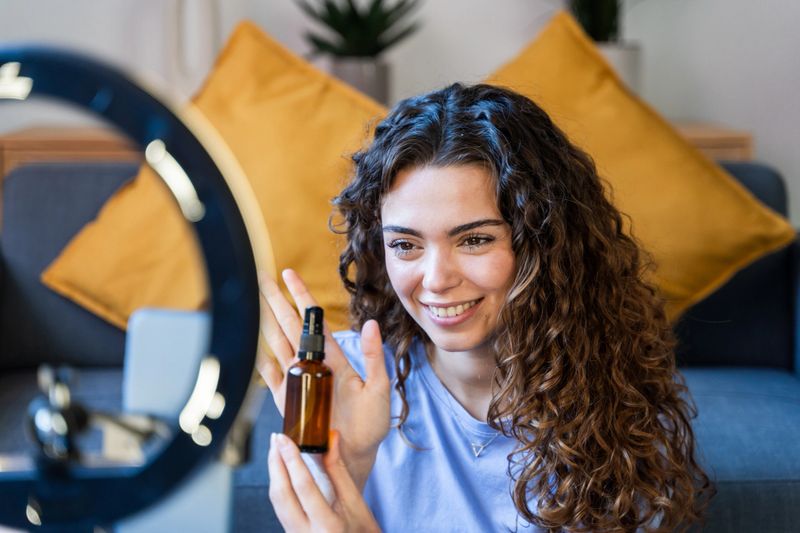 Curly-haired woman showing a skincare oil bottle while recording a video with a laptop and ring light