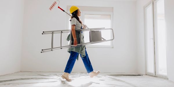 Woman in a yellow hard hat carrying a ladder and paint roller in an empty room.