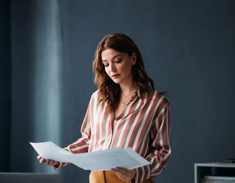 Focused young professional woman intently reviewing papers, dressed in a chic striped blouse in a modern workspace.
