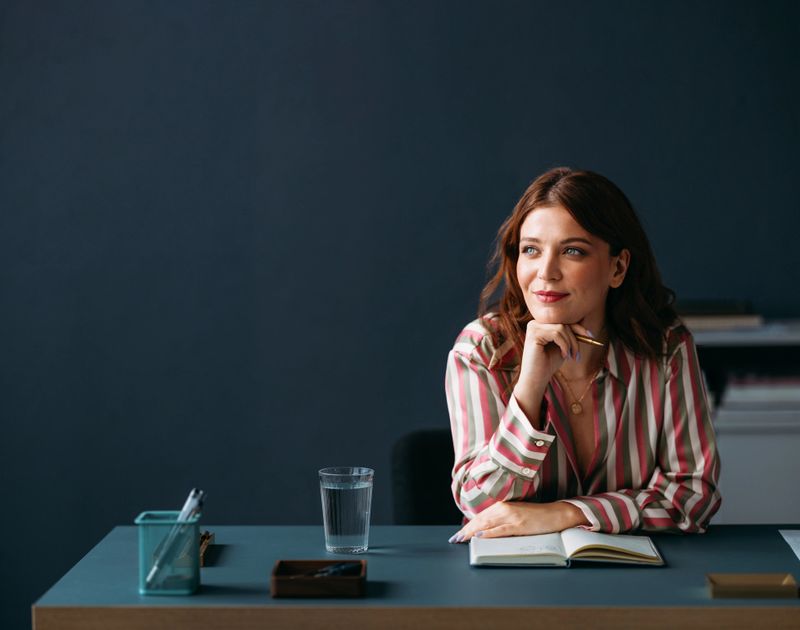 Confident businesswoman with a thoughtful smile sitting at her desk in a modern office setting, evoking professionalism and contentment.