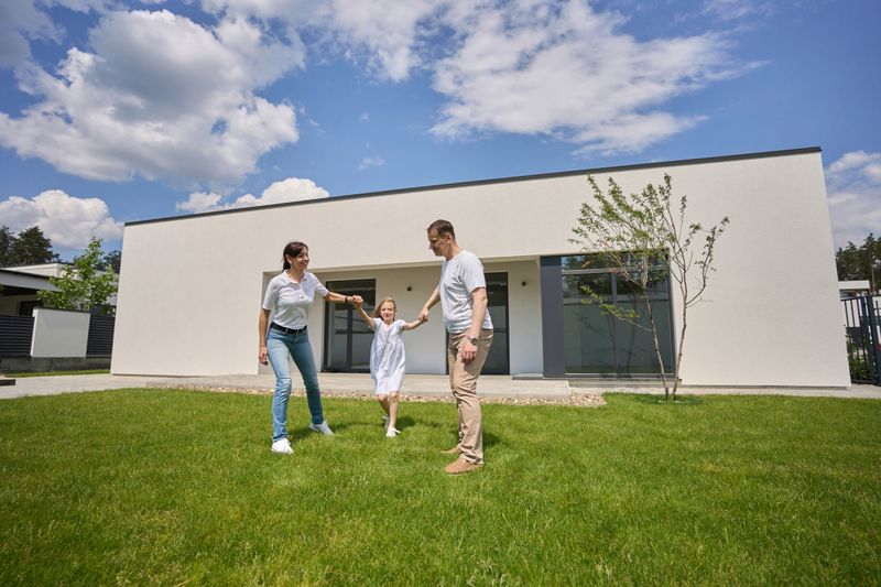 Pleased caucasian mother, father and little daughter holding hands on green yard near modern townhouse in warm sunny day. Happy family relationship and spending time together
