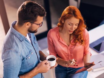 Two coworkers discuss work while one holds a coffee cup and the other uses a tablet.