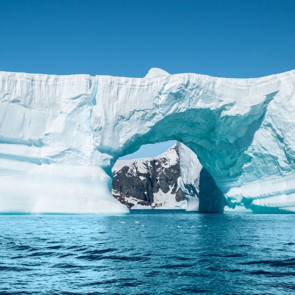 A large ice arch over blue water with snow-covered mountains in the background.