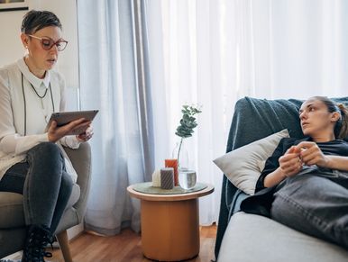 A woman therapy patient lies on a couch while a therapist takes notes on a tablet.