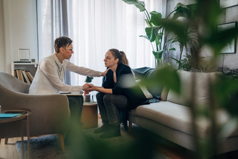 A Therapist is seen offering comfort to Young woman who appears to be in distress and cry. They are seated closely on a couch, creating an atmosphere of support and understanding.