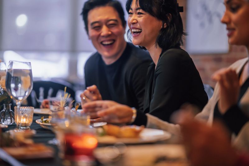 A large group of adults sit together in a restaurant as they enjoy a meal together.  They are each dressed semi-formally and are smiling and laughing as they enjoy the dinner party and their food.