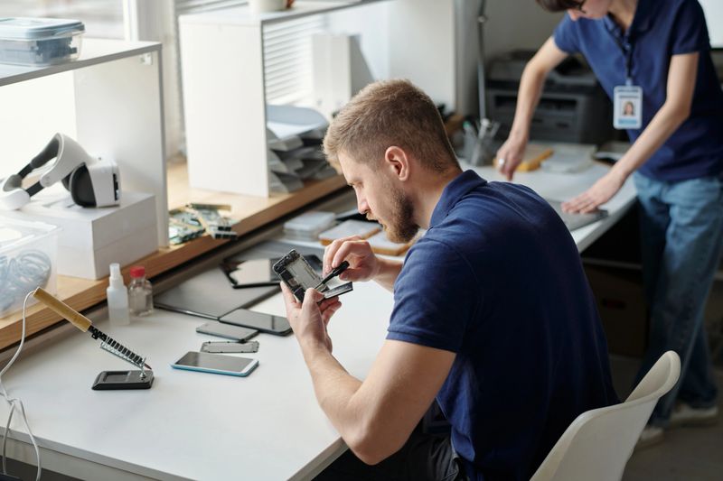 Young worker of technical repair office fixing tiny details of disassembled cellphone with small screwdriver while sitting by workplace
