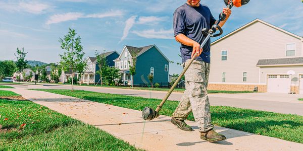 Man trimming grass along a sidewalk in a suburban neighborhood.