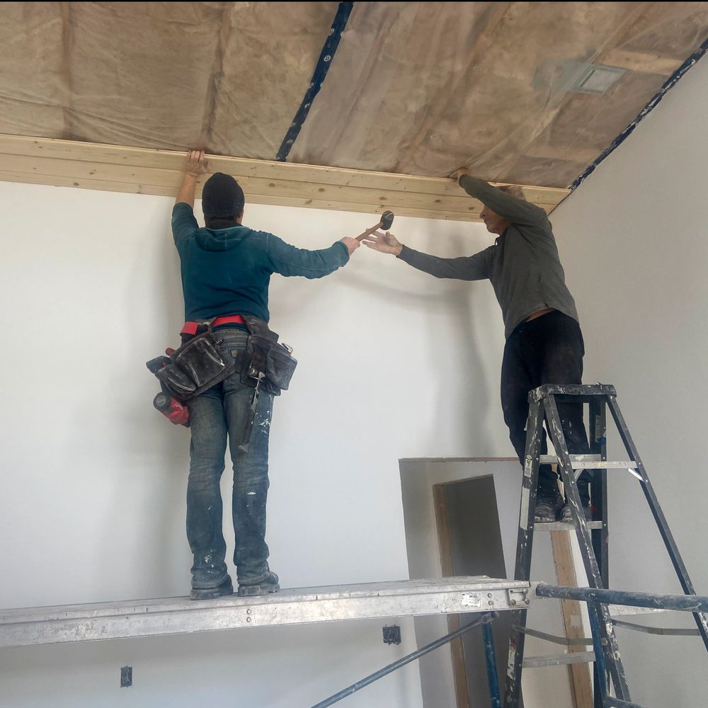 Two workers installing wood paneling on a ceiling inside a room.