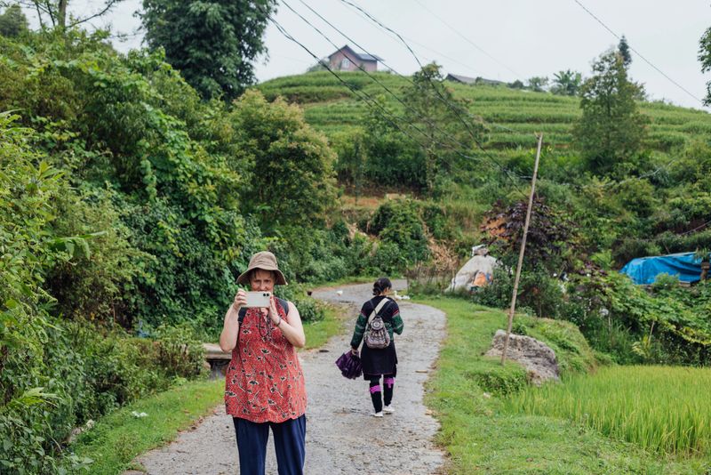 A hill tribe lady guide dressed in traditional dress holding a purple umbrella is guiding a European tourist through a curved path amongst paddy fields and vegetable plots in the area of Sa Pa, Vietnam. The tourist has turned to the camera, to take a photograph with her phone. This authentic shot captures the reality of the area.