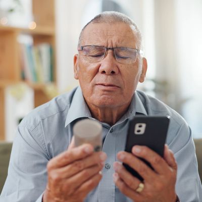 Elderly man struggling to read medication label while holding phone.