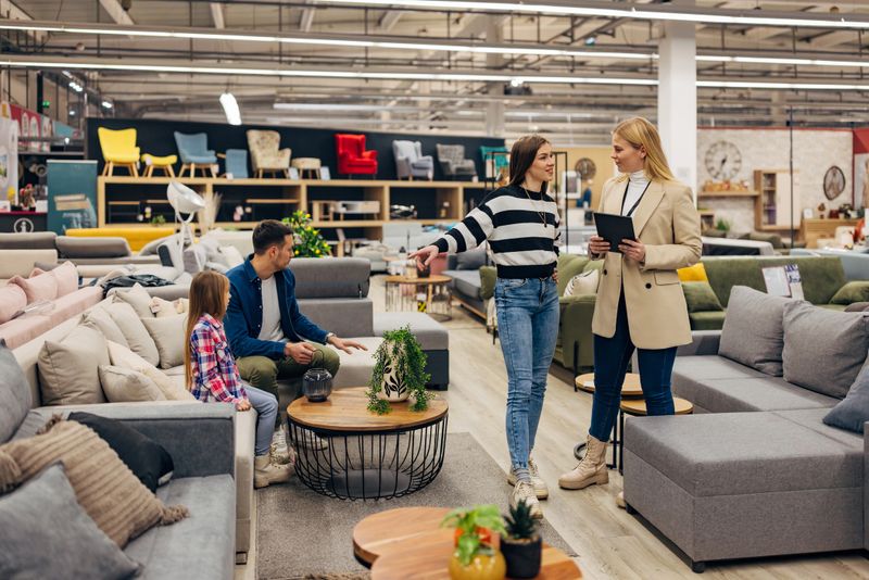 Carefree saleswoman uses a digital tablet to show the smiling female customer an offer of different sofa beds that they have in stock