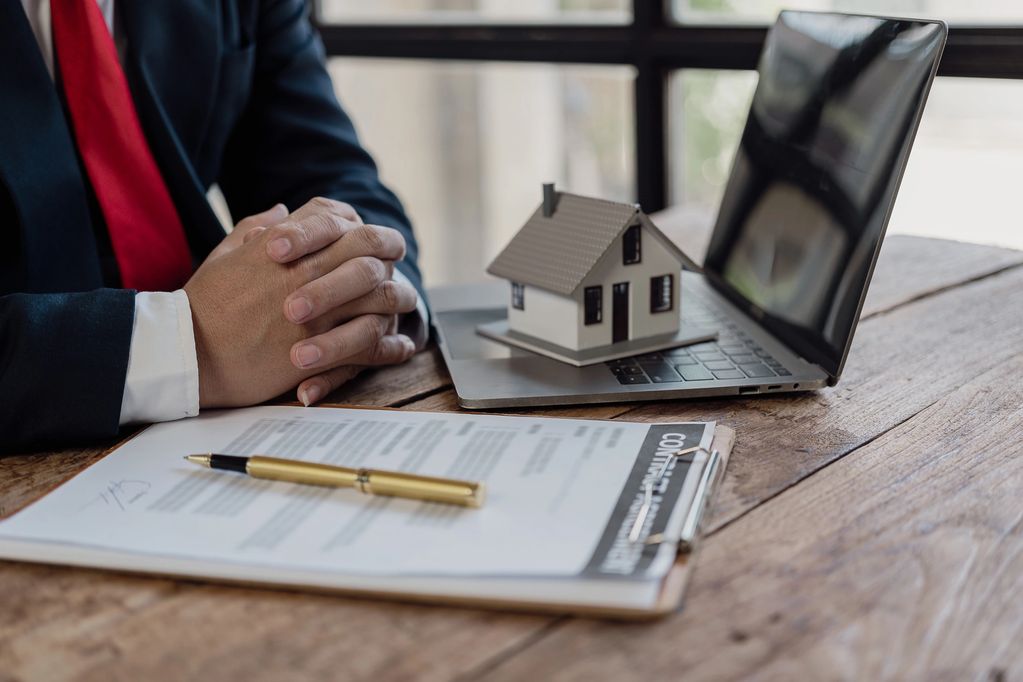 a person sitting at a table with a laptop, and a note pad. With a miniature house on the laptop keys