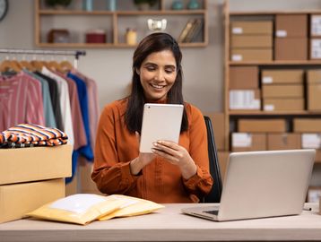 Woman using a tablet in a workspace with packages and clothing rack.