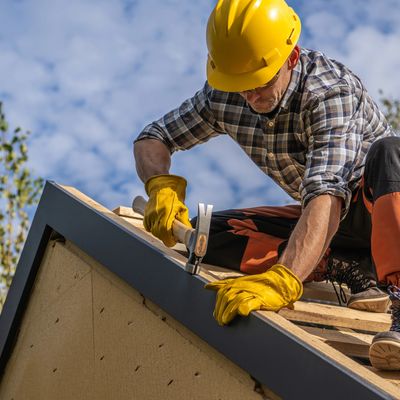 Construction worker wearing safety gear hammers nails into roof framing under a partly cloudy sky.