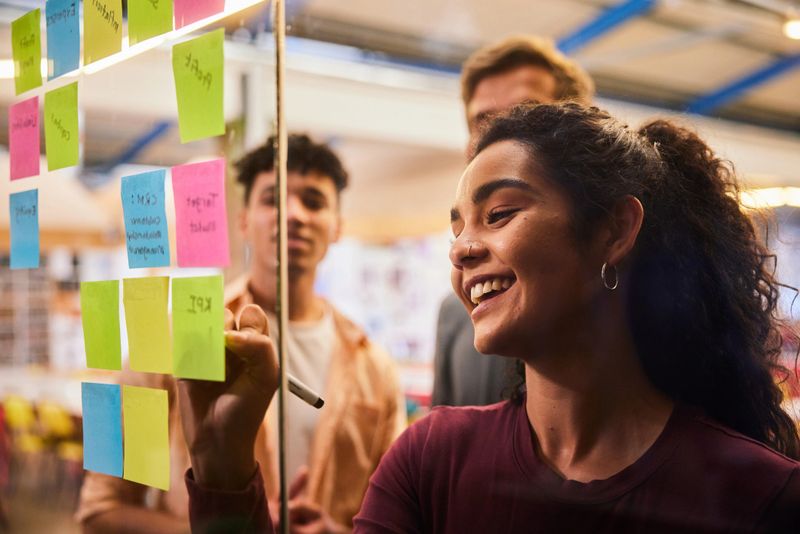 Smiling young female college student and some male classmates brainstorming together with adhesive notes on a glass wall in a classroom