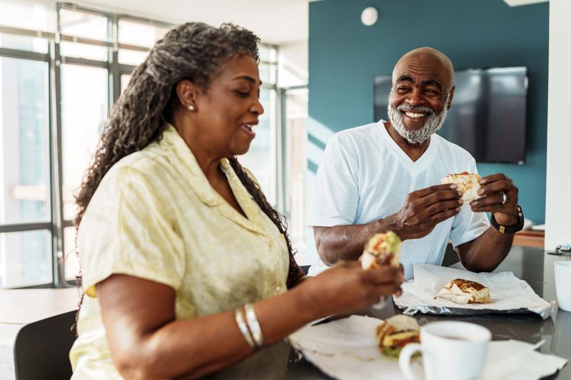 A vibrant senior couple of African American descent wearing pajamas eats breakfast burritos they ordered from room service in their luxury hotel while spending a relaxing weekend exploring Washington DC.