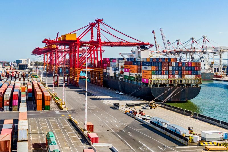 Elevated view container ship loading/unloading Port of Melbourne Swanson Dock container terminal. Three moored container ships are being loaded/unloaded by a row of red gantry cranes on the left with a row of white cranes on the right of frame. Also to the left are rows of multi-coloured stacked containers waiting for transportation. Multiple logos visible, none dominant, distinctive ID and ship/vessel names edited.
