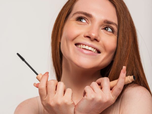 Smiling woman holding mascara and foundation makeup products.