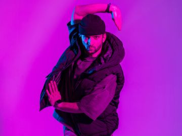 Man in a black hat and vest striking a dynamic pose under purple lighting.