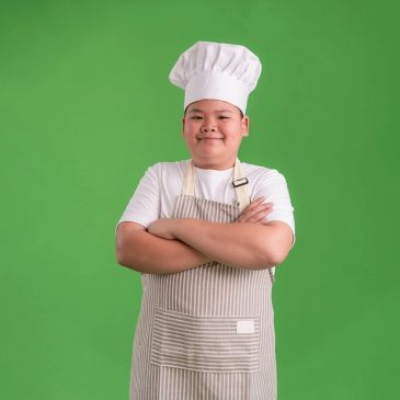 Confident young chef in white apron and hat with arms crossed against green background.