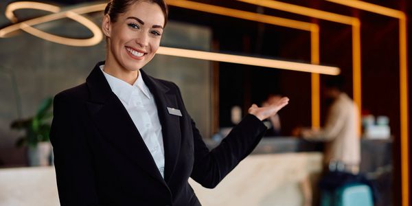 Smiling hotel receptionist welcoming guests with an inviting gesture at the front desk.