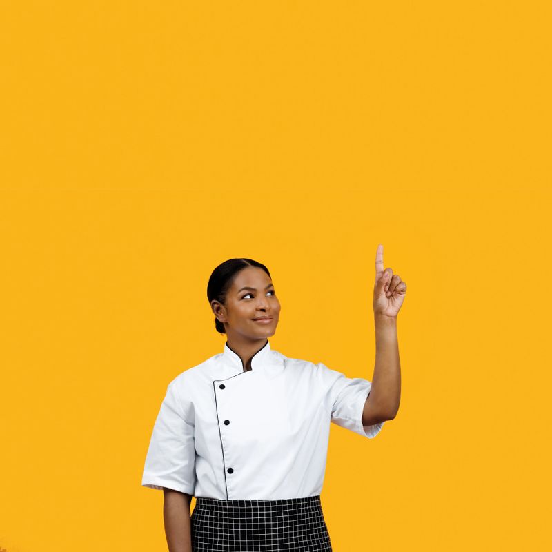 Cheerful black female chef in white jacket pointing upward at copy space above her head, hinting at new idea or concept, posing against bright yellow background, illustrating creativity