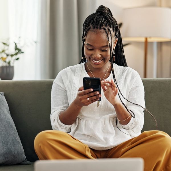 A smiling woman using her smartphone while sitting cross-legged on a couch.