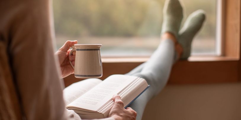 Person relaxing by a window, reading a book and holding a mug.