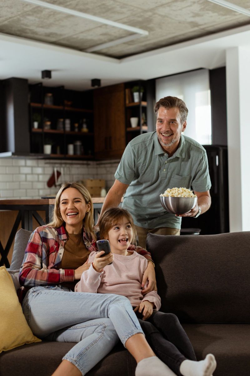 A family of three is comfortably nestled on a couch, their faces reflecting excitement and attentiveness as they share a bowl of popcorn during a suspenseful movie night
