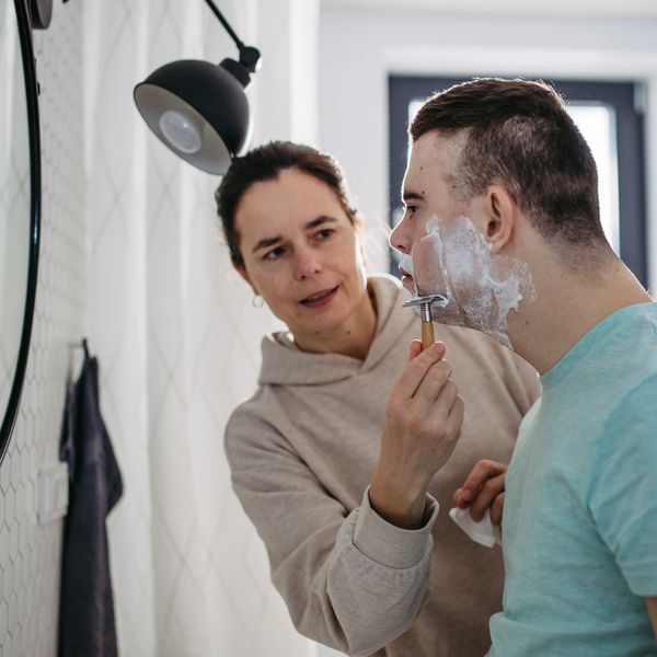 A woman helps a young man shave his face in a bathroom mirror.
