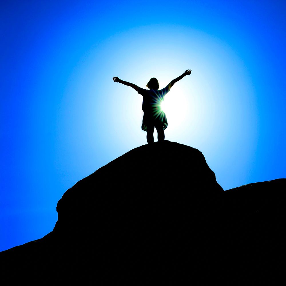 Silhouette of a person standing triumphantly on a hill against a bright blue sky.