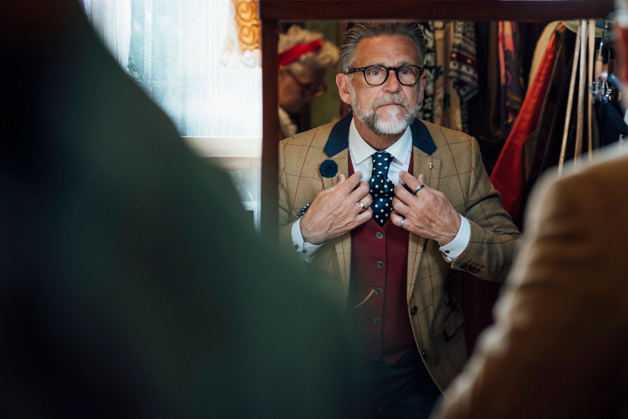 An elderly man adjusts his tie in front of a mirror, dressed in a stylish suit.