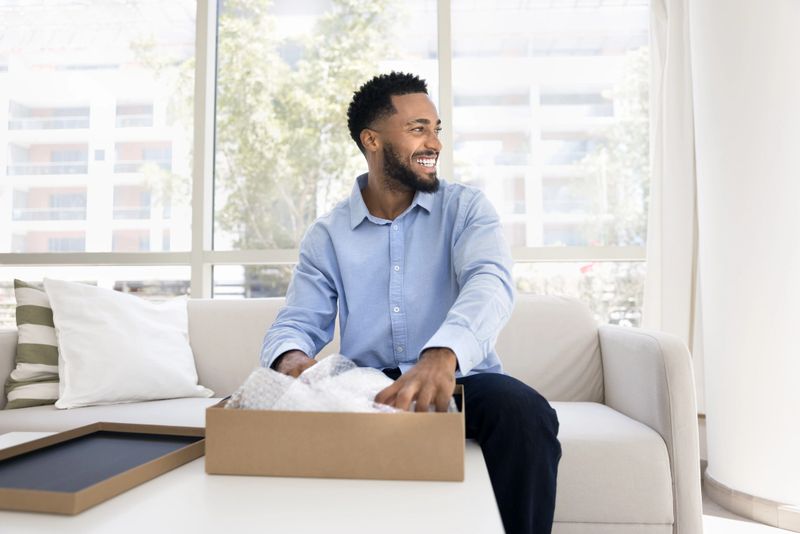 Positive young African American customer guy unpacking parcel at home, opening cardboard box with purchase from online store, looking away with toothy smile, laughing, thinking