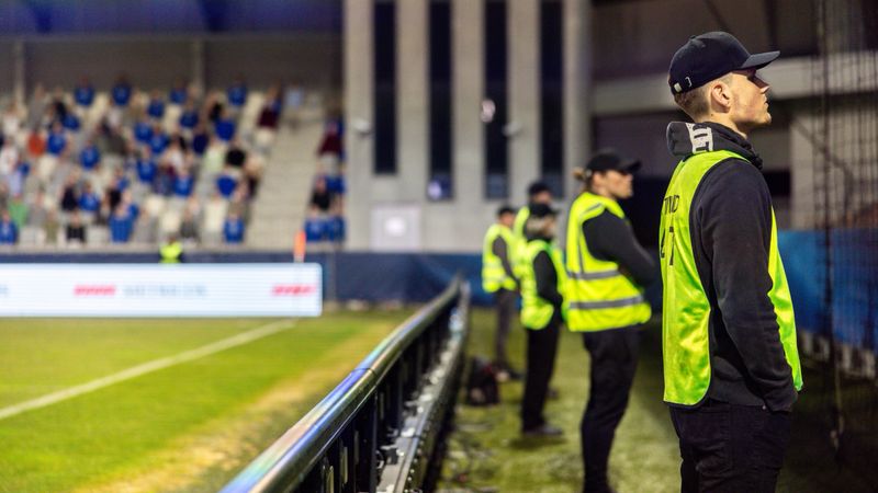 Male security personnel in high-visibility jackets maintain vigilance during a nighttime football match. Professional attire and the outdoor stadium setting underscore the safety measures in place.