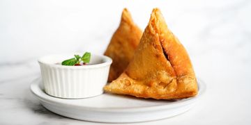 Two golden samosas served with a white bowl of chutney on a white plate.