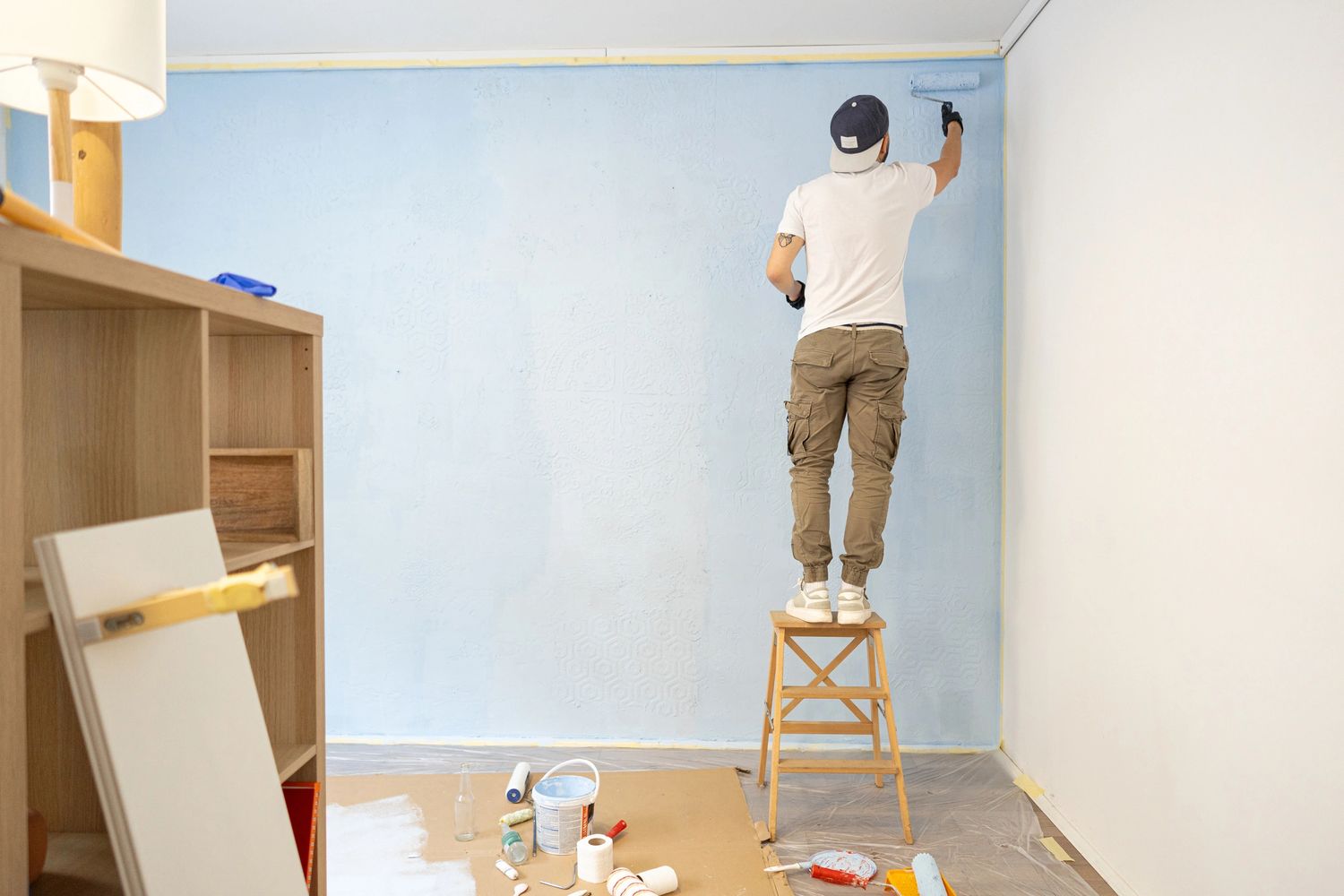 Person painting a wall light blue while standing on a wooden stool.